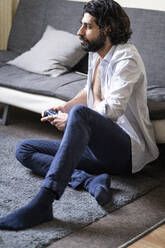 Young man playing video game while sitting in front of sofa at home - FBAF01629