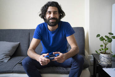 Smiling man with game controller sitting on sofa at home - FBAF01630