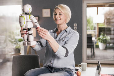Smiling businesswoman holding model of robot while sitting on table at home office - UUF23085