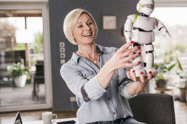 Fröhliche Unternehmerin, die bei der Arbeit zu Hause ein Robotermodell untersucht - UUF23086