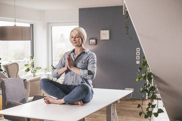 Lächelnde Frau mit geschlossenen Augen übt Yoga auf dem Tisch zu Hause - UUF23090