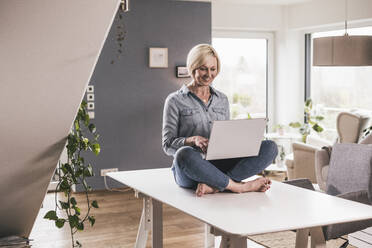 Smiling businesswoman working on laptop while sitting on table at home office - UUF23092