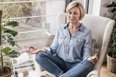Frau mit geschlossenen Augen sitzt im Lotussitz auf einem Sessel zu Hause - UUF23097