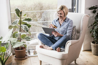 Lächelnde Frau, die einen digitalen Tisch benutzt, während sie Essen im Wohnzimmer hält - UUF23098