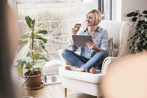 Lächelnde Frau mit digitalem Tablet, die einen Donut betrachtet, während sie zu Hause auf einem Sessel sitzt - UUF23099