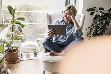 Kontemplative Frau mit digitalem Tablet, die im Wohnzimmer wegschaut - UUF23100