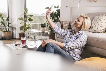 Lächelnde reife Frau, die ein Selfie macht, während sie sich auf das Sofa im Wohnzimmer lehnt - UUF23104