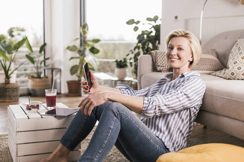 Smiling woman with smart phone sitting on floor in living room - UUF23105
