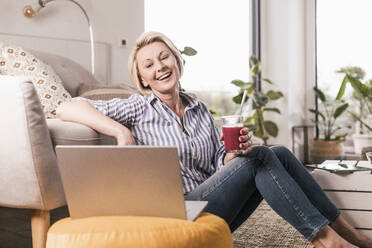 Glückliche reife Frau, die ein Saftglas hält, während sie zu Hause auf den Laptop schaut - UUF23106