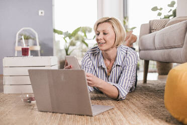 Blonde Frau mit Laptop und Smartphone, während sie zu Hause liegt - UUF23108