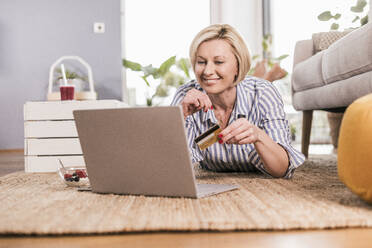 Blonde reife Frau mit Kreditkarte beim Online-Shopping mit Laptop zu Hause - UUF23110