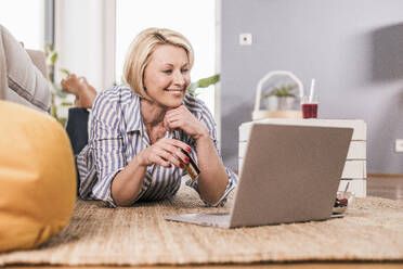 Smiling woman with credit card doing online shopping while lying on front at home - UUF23112