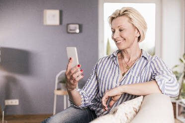 Lächelnde reife Frau, die ein Mobiltelefon benutzt, während sie zu Hause auf dem Sofa sitzt - UUF23114