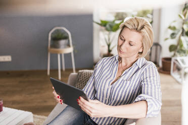 Mature woman using digital tablet while sitting on sofa in living room - UUF23118