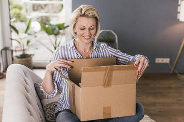 Lächelnde reife Frau beim Öffnen einer Schachtel zu Hause - UUF23119