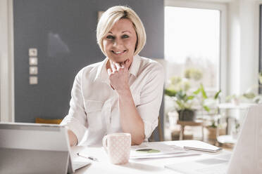 Smiling female entrepreneur with digital tablet and laptop sitting at table in home office - UUF23122