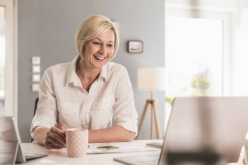 Lächelnde Geschäftsfrau bei einem Videogespräch über den Laptop, während sie zu Hause Kaffee trinkt - UUF23124