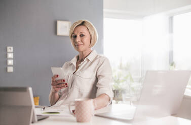 Smiling businesswoman with smart phone in home office - UUF23127