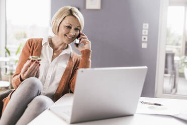 Lächelnde Unternehmerin, die mit ihrem Smartphone telefoniert und etwas isst, während sie zu Hause arbeitet - UUF23148