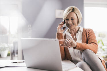 Smiling female professional pointing at laptop while communicating through smart phone at home - UUF23150