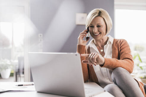 Lächelnde weibliche Fachkraft, die auf einen Laptop zeigt, während sie über ein Smartphone zu Hause kommuniziert - UUF23150