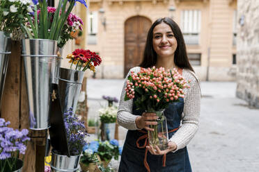 Young female florist holding Hypericum flower vase while standing at flower shop - EGAF02111