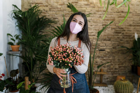 Junge weibliche Floristin mit Gesichtsschutzmaske hält Hypercium Blumenvase im Geschäft - EGAF02112