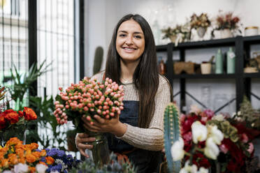 Lächelnde Blumenhändlerin mit einer Vase voller Blumen im Laden stehend - EGAF02117