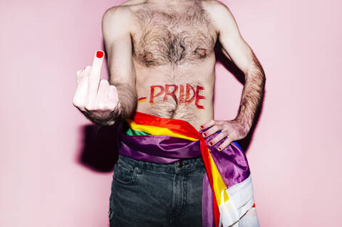 Gay man with rainbow flag tied around waist showing middle finger standing against pink background stock photo