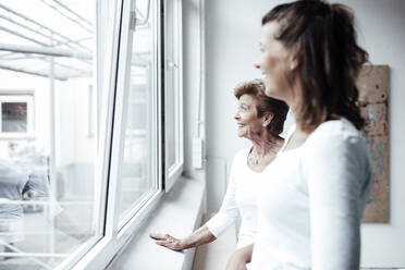 Glückliche Großmutter mit Enkelin, die durch das Fenster ihres Hauses schaut - GUSF05472