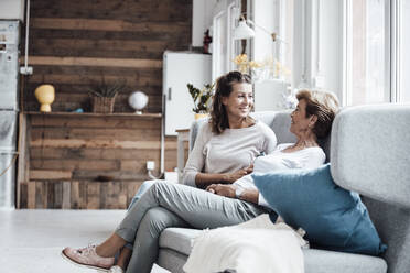 Smiling young woman talking with grandmother while relaxing on sofa at home - GUSF05496