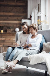 Junge Frau, die ein digitales Tablet benutzt, während sie mit ihrer Großmutter auf dem Sofa zu Hause sitzt - GUSF05500