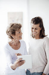 Aufgeregte ältere Frau, die ein digitales Tablet hält, während sie sich mit ihrer Enkelin zu Hause unterhält - GUSF05508