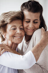 Lächelnde Großmutter mit geschlossenen Augen, die eine junge Frau zu Hause umarmt - GUSF05510