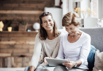 Lächelnde Frau schaut auf ein digitales Tablet, während sie bei ihrer Enkelin zu Hause sitzt - GUSF05520