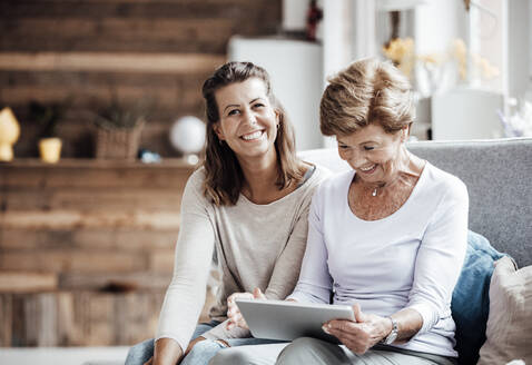 Lächelnde Frau schaut auf ein digitales Tablet, während sie bei ihrer Enkelin zu Hause sitzt - GUSF05520