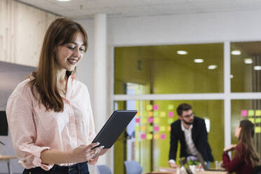 Smiling young businesswoman working on digital tablet with colleagues in background at workplace - PNAF01122