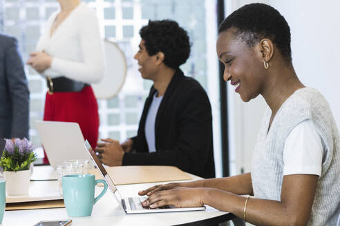 Lächelnde Geschäftsfrau arbeitet am Laptop mit Kollegen im Hintergrund im Büro - PNAF01149