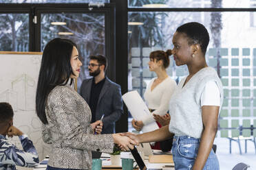 Female entrepreneurs shaking hands while standing with colleagues in background at board room - PNAF01152