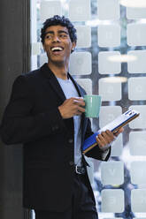 Lächelnder Geschäftsmann mit Kaffeetasse und Aktenordner, der am Fenster steht - PNAF01194