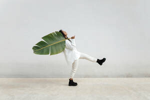 Junge Frau tanzt, während sie ein großes Bananenblatt an einer weißen Wand trägt - TCEF01707