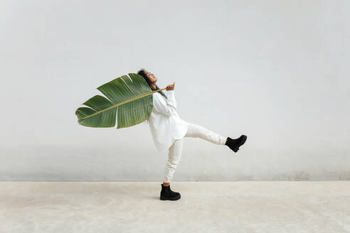 Young woman dancing while carrying big banana leaf by white wall - TCEF01707