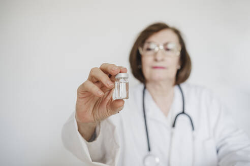 Senior female doctor holding vial in front of white wall - EBBF02798