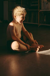 Happy boy sitting on floor at home during dusk - MFF07670