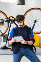 Mann, der ein digitales Tablet benutzt, während er sich zu Hause auf einen Tisch mit einem umgestürzten Fahrrad stützt - GIOF11928