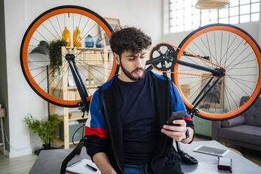 Junger Mann, der ein Mobiltelefon benutzt, während er sich zu Hause mit dem Fahrrad auf einen Tisch stützt - GIOF11933