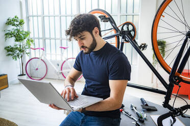 Young man using laptop while leaning on table by bicycle at home - GIOF11950