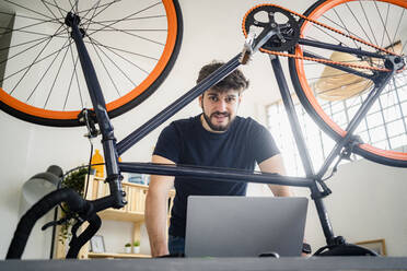 Mann, der einen Laptop benutzt, während er sich zu Hause auf einen Tisch mit umgestürztem Fahrrad stützt - GIOF11955