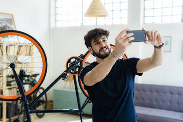 Junger Mann, der ein Selfie mit seinem Smartphone macht, während er zu Hause in der Nähe eines umgestürzten Fahrrads steht - GIOF11963