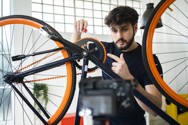 Männlicher Vlogger, der auf die Fahrradkette zeigt, während er ein Tutorial zu Hause filmt - GIOF11965